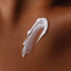 Close-up texture shot of Natural Moisturizing Factors + Inulin Body Lotion. The white, creamy product is swatched on smooth, deeply toned skin to show its rich, moisturizing consistency.